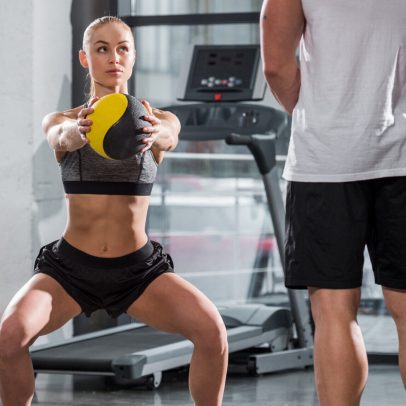 back view of personal trainer watching at sportswoman training with fitness ball in gym