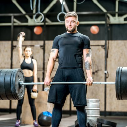 Handsome athletic man in black sports wear lifting up a heavy burbell with woman training on the background in the gym